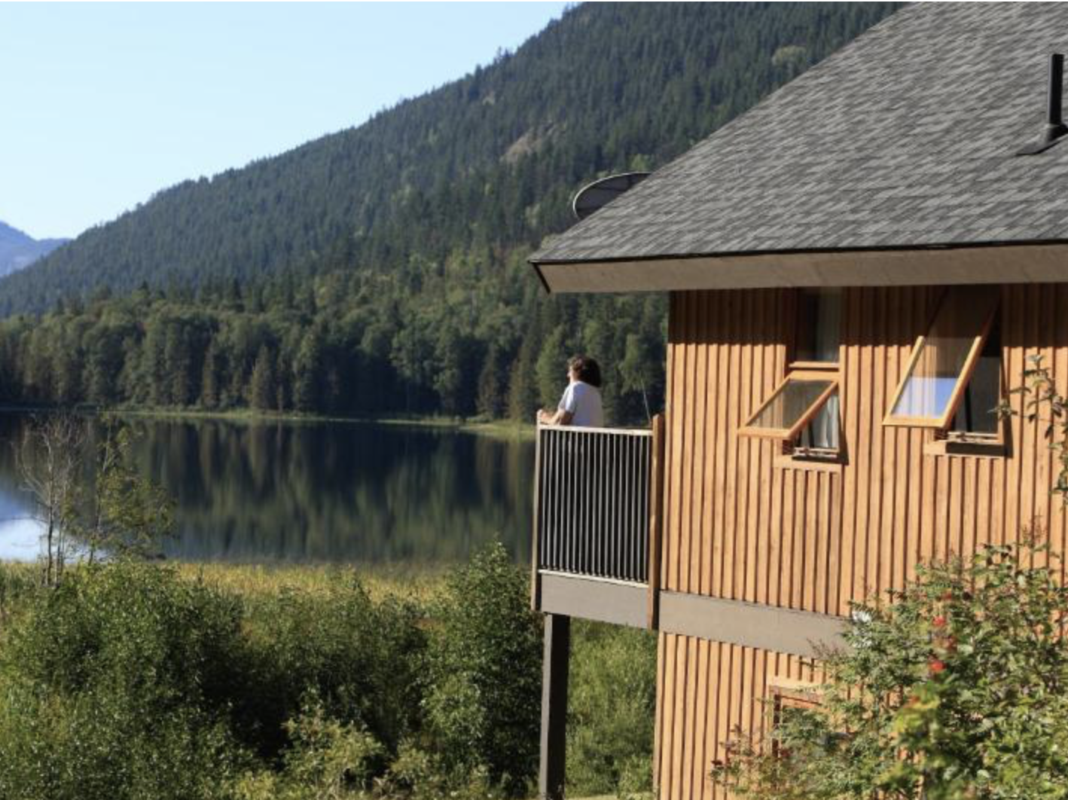 Wooden lakeside cabin with a balcony; a person stands at the railing looking over the calm lake and forested hills nearby.