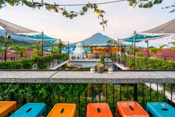 Rooftop bar terrace with string lights, shade sails, and lush greenery, a mountain in the background; colorful orange and blue bins line the railing in the foreground.