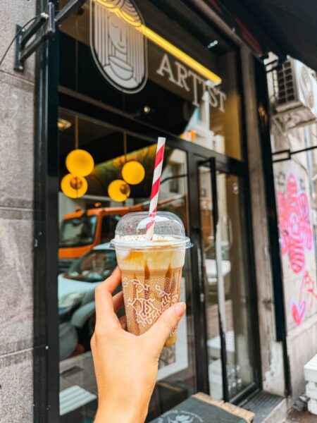 Hand holding a clear cup with iced coffee and a red-striped straw outside a cafe storefront