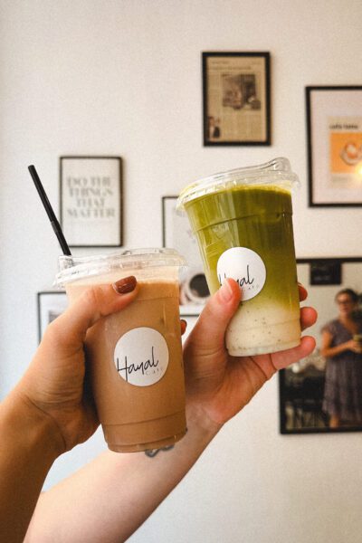 Two hands hold up two takeaway cups, one green smoothie and one iced coffee, in a cafe with framed wall art behind.