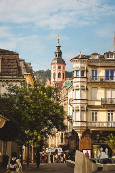 Street lined with ornate pastel buildings; a pink church tower rises in the background as pedestrians walk along the shaded avenue.