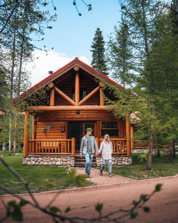 Couple walking hand in hand on a path in front of a wooden log cabin surrounded by tall trees on a sunny day.