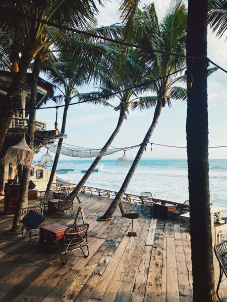 Beachside wooden deck cafe with palm trees, wicker chairs, and an ocean view behind.