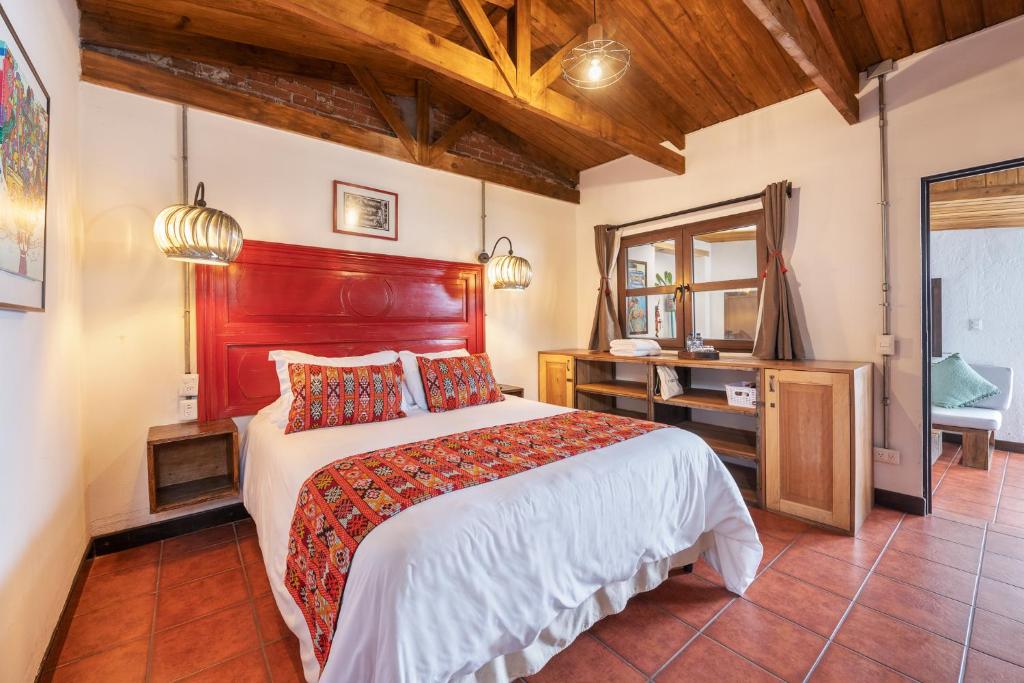 Cozy rustic hotel bedroom with a bright red headboard, white bedding, and a colorful patterned runner with pillows on a tiled floor. Wooden ceiling beams overhead.