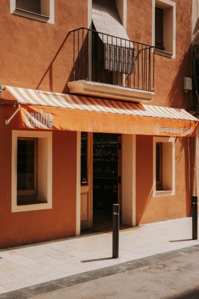 Orange stucco storefront with a striped orange-and-white awning, open doorway, and windows above; balcony with black railing on the second floor.
