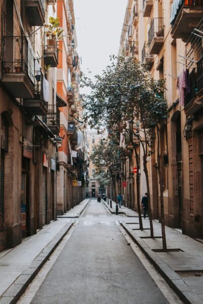 Narrow European street between tall peach-colored buildings with balconies, laundry hanging, and a tree in the center; pedestrians in the distance.