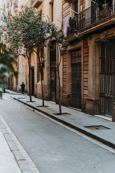 Narrow city street lined with trees and beige buildings; laundry hanging from balconies and a person walking in the distance.