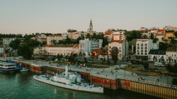Riverfront promenade with a white ship docked at the quay, people walking along the seawall and a row of colorful buildings behind.