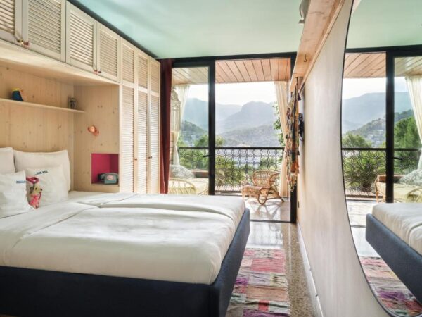 Hotel room with a large bed, built-in wood shelves, and a balcony with mountain views through glass doors.