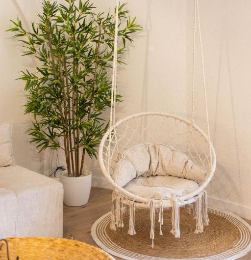 Beige hanging wicker chair with cushions in a light, cozy living corner beside a tall potted plant and round woven rug.