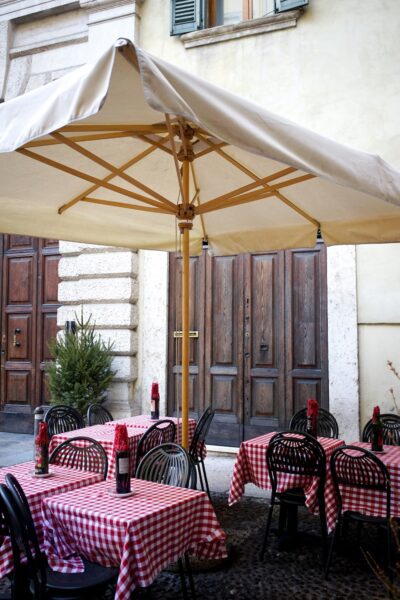 Outdoor cafe seating: red-and-white checkered tables under a large beige umbrella on a cobblestone street, with dark metal chairs and wooden doors in the background.