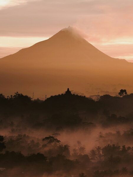 Silhouetted volcanic peak at dawn with a misty tropical forest in the foreground and a small temple on a hilltop.
