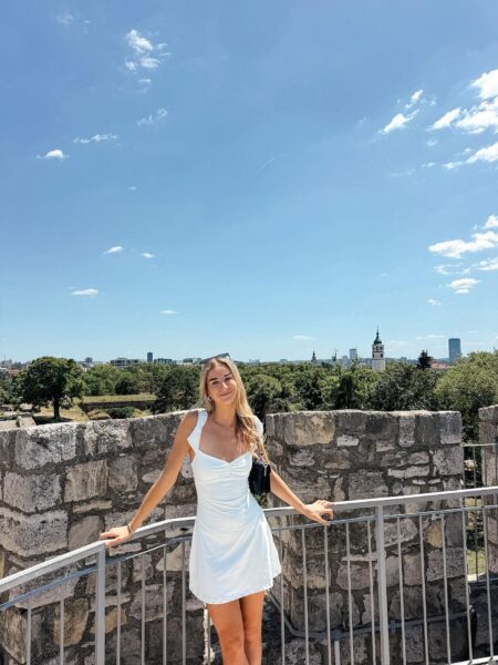 Woman in a white dress posing at a stone railing on a sunny day, with a city skyline and blue sky behind her.