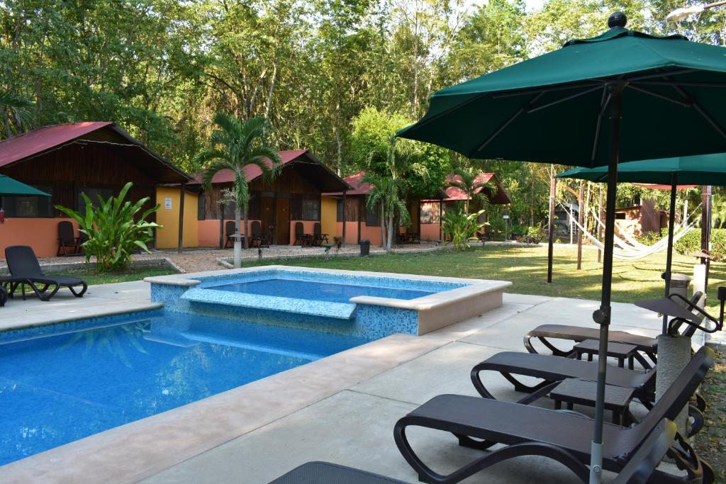 Resort pool area with blue tiled pool, shallow spa, and lounge chairs under large umbrellas amid tropical plants and huts.