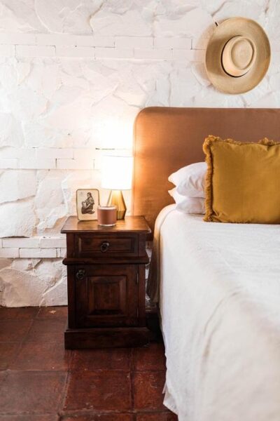 Cozy bedroom corner with a dark wooden nightstand, lamp, framed photo, and a hat on the white brick wall above a bed with a mustard pillow and white linens