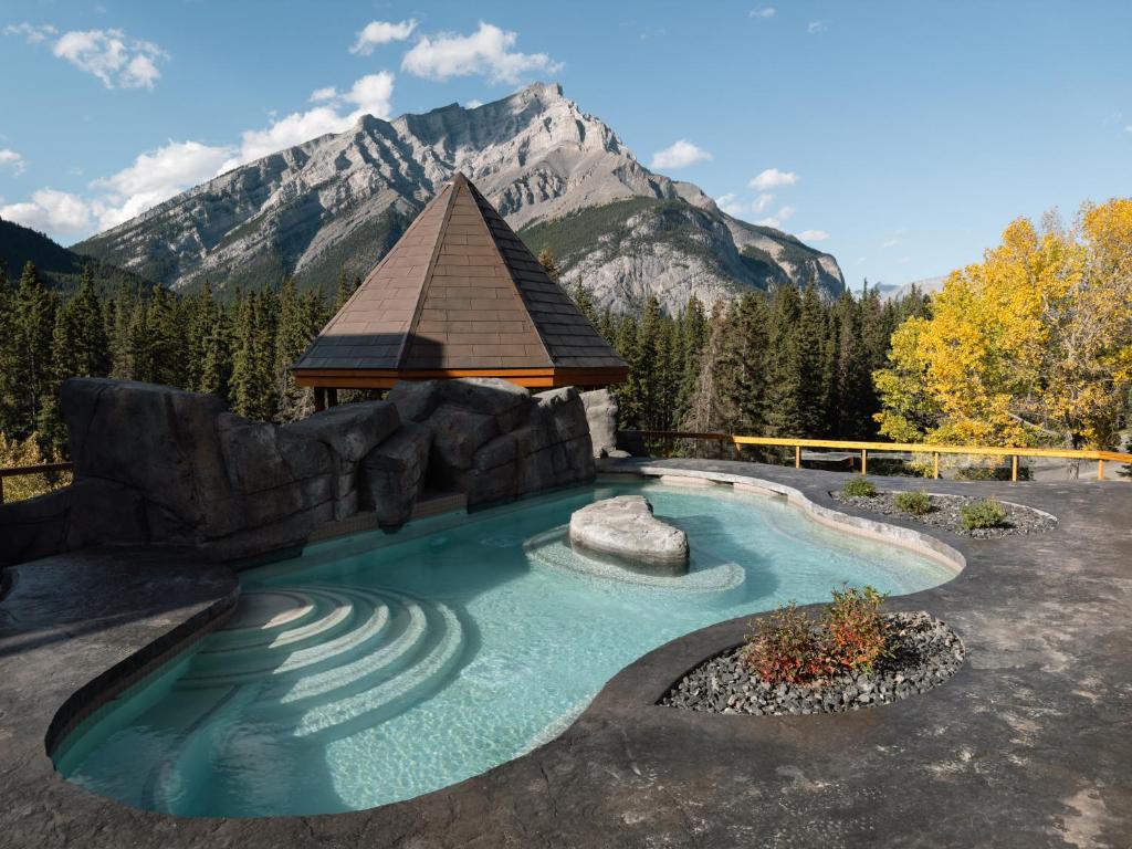 Outdoor pool with rock features and a wooden pyramid-roof structure, set against a forest and mountain backdrop on a sunny day.