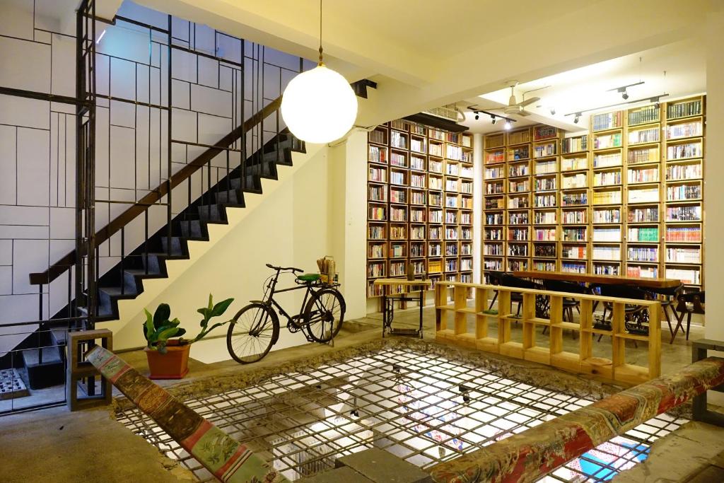 Cozy library interior with tall bookshelves, a staircase, and a bicycle leaning by a potted plant near a glass-floor feature under warm lighting.