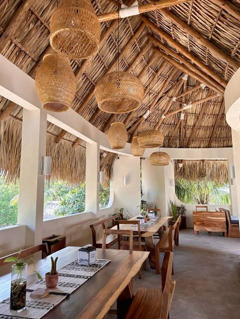 Open-air restaurant with a thatched roof, woven pendant lights, wooden tables and chairs, and greenery outside.