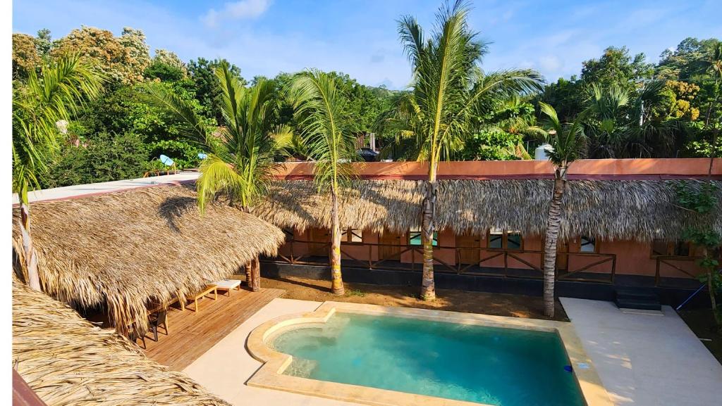 Resort courtyard with thatched-roof huts surrounding a small pool and palm trees in a sunny tropical setting.