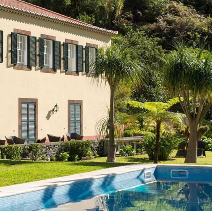 Two-story beige villa with green shutters, outdoor seating along a hedge, and a blue-tiled pool in the foreground. Palm trees and tropical plants surround the scene.