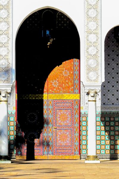 Ornate arched doorway with vibrant geometric tile panels in orange, pink, and teal, framed by white patterned walls. A dark silhouette stands in front of the arch.