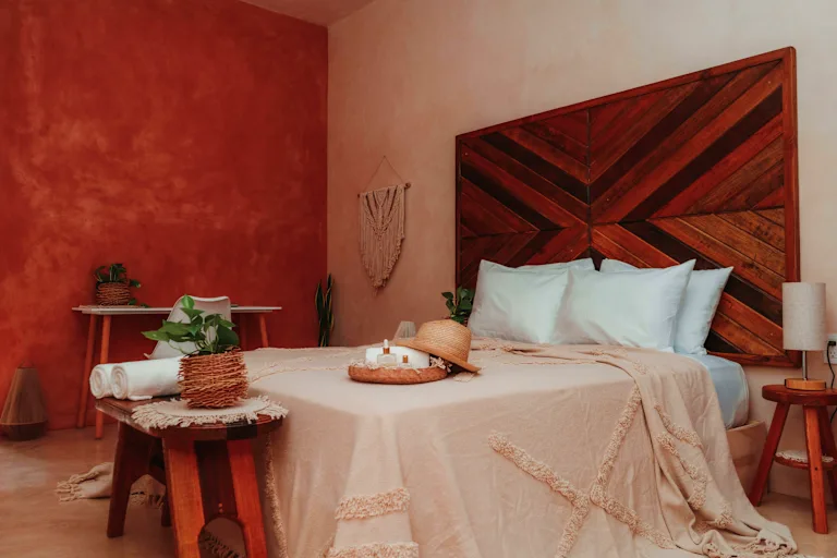 Cozy bedroom with a large geometric wooden headboard, warm terracotta wall, and a bed with beige throw and light-blue pillows.