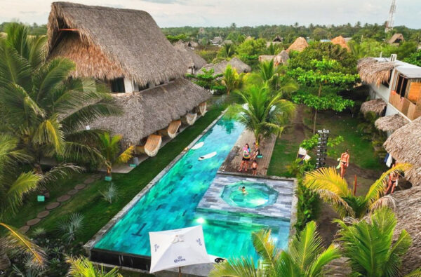 A tropical resort courtyard with thatched-roof cottages, a long turquoise pool, a circular hot tub, and palm trees with guests relaxing nearby.