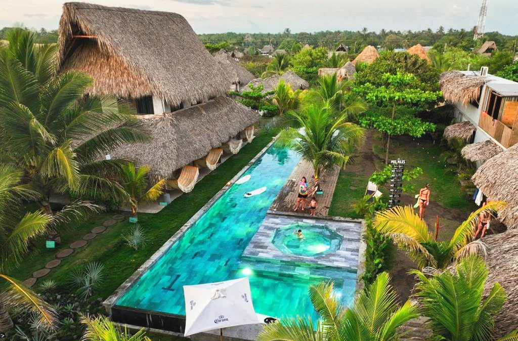 A tropical resort courtyard with thatched-roof cottages, a long turquoise pool, a circular hot tub, and palm trees with guests relaxing nearby.