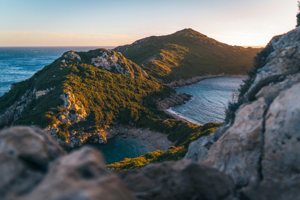 Coastal landscape showing forested hills along a rocky coastline with a small sandy beach in a crescent bay at sunset, ocean on the left.