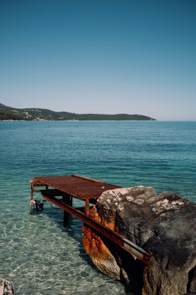 Rusty metal platform attached to rocks at a turquoise sea shore with hills in the distance.