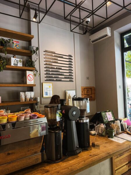 Cafe counter with two espresso machines, colorful cups, and a wall-mounted menu board listing drinks like Espresso, Cappuccino, Latte.