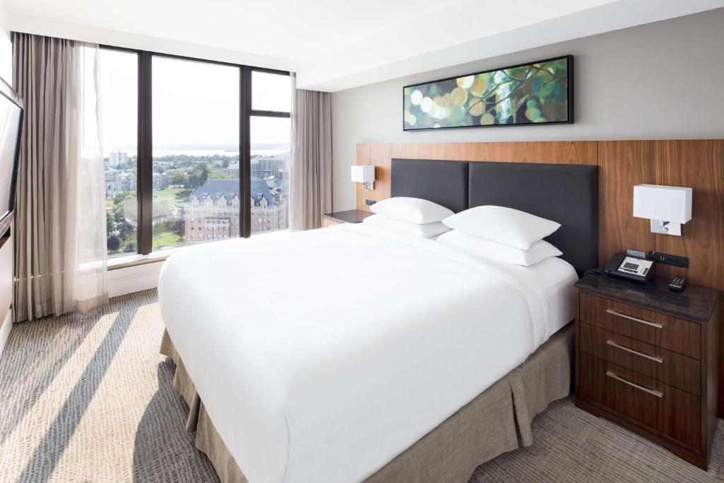 Hotel room with a large white bed, two nightstands, and a city view through tall windows.