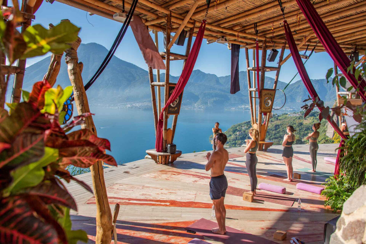 Group of people practicing yoga on a wooden deck with a lake and mountains in the background beneath a bamboo canopy.