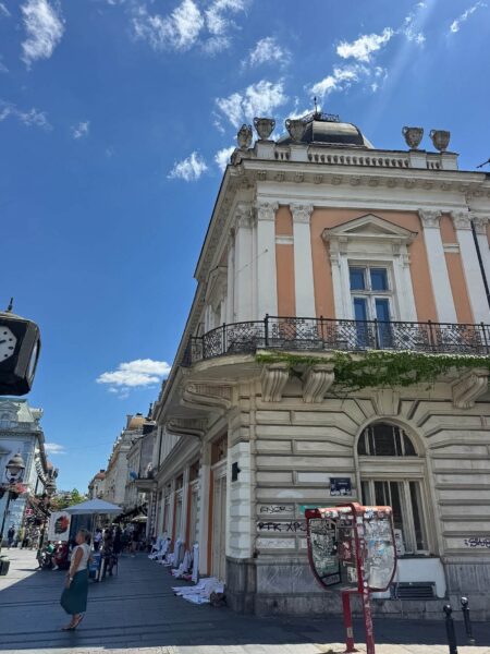 Corner of a historic peach-and-white building with ornate columns, a black wrought-iron balcony, and a bright blue sky above.