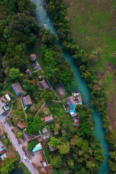 Aerial view of a turquoise river winding between dense green forests and a small village with thatched and tiled huts.