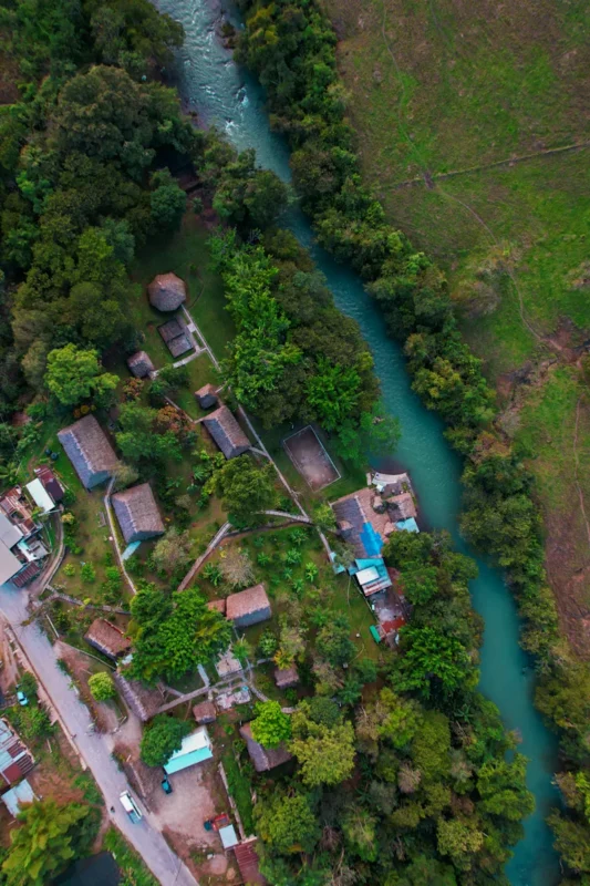 Aerial view of a turquoise river winding between dense green forests and a small village with thatched and tiled huts.