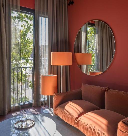 Cozy living room corner with an orange sofa, matching floor lamp, and a round wall mirror on a red accent wall; sheer curtains shade a balcony with trees outside, sunlight casting shadows on the coffee table.