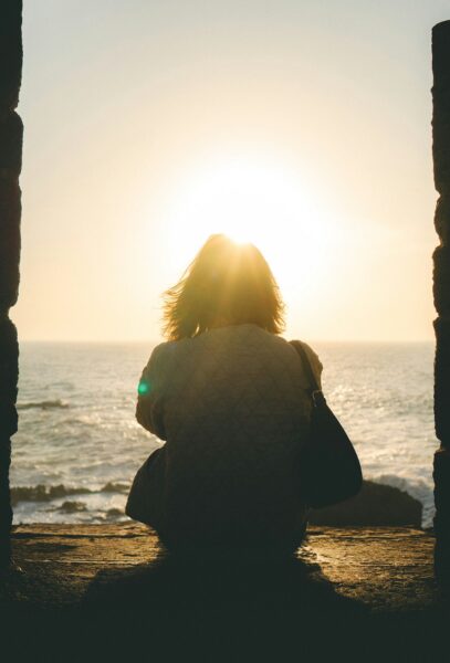 Person sitting on a seawall at sunset, facing the ocean with the sun behind them and a bag on their shoulder