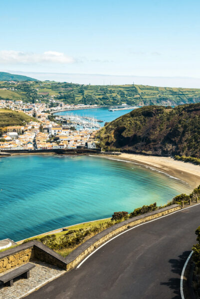 Coastal town along a turquoise bay with a marina, terraced hills, and a winding road along the coastline.