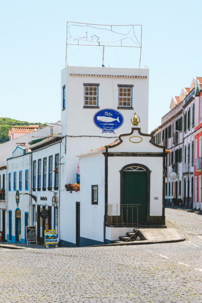 White corner building with blue shop sign 'Peter Café Sport' and a small chapel-like entrance, cobblestone street.