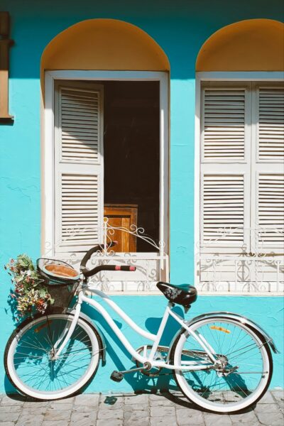 White vintage bicycle with a flower-filled basket parked against a bright turquoise wall with yellow arches above a window.