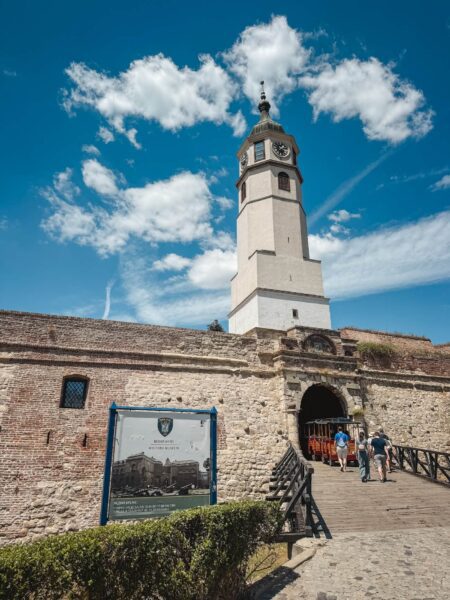 Clock tower rising above a stone fortress wall on a bright day, with visitors approaching the entrance path beneath a blue sky.