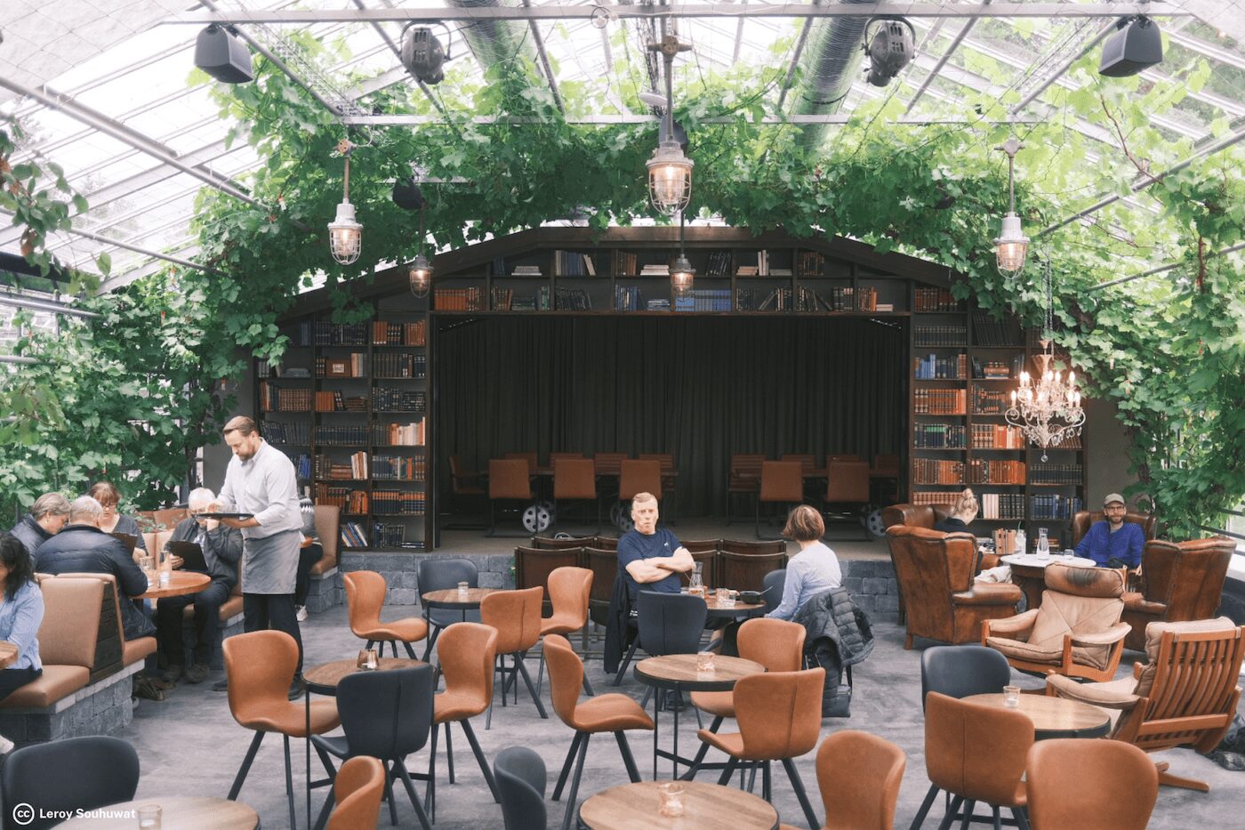 Cafe inside a glass greenhouse with ivy-covered ceiling, bookshelves along the walls, and a small stage at the back surrounded by tables and chairs.