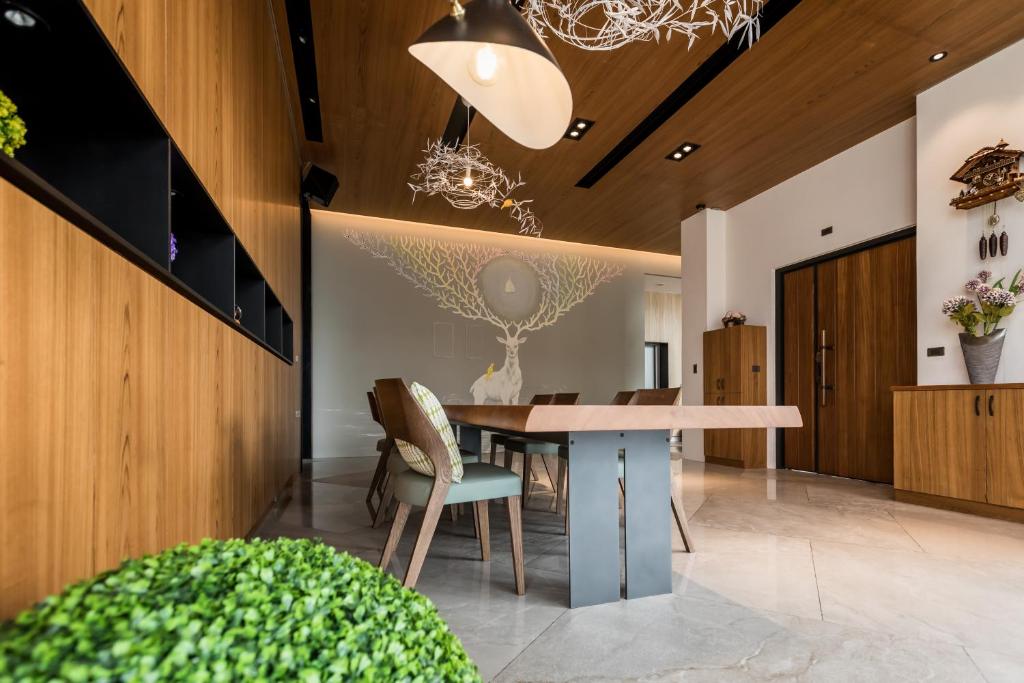 Modern dining room with a long wooden table, teal chairs, and a wooden paneled wall; a sculptural chandelier hangs from a wood ceiling, and a tree mural decorates the back wall.