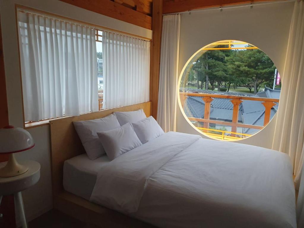 Bedroom with a large round window, white bedding, and sheer curtains; warm wooden beams and a small bedside lamp on the left.