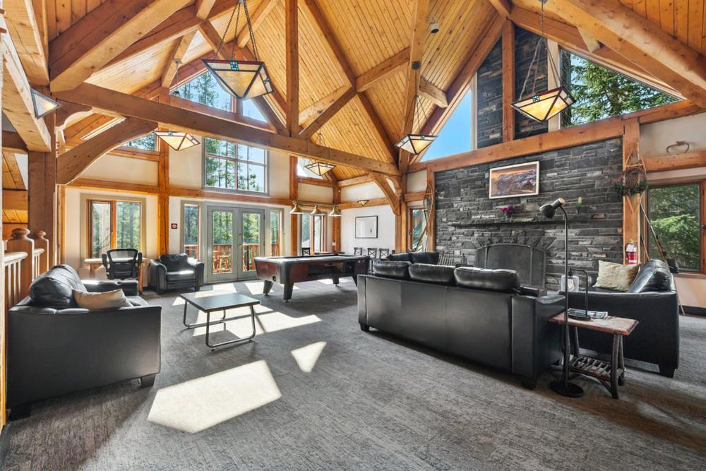 Open living space with exposed wooden beams, large windows, and a stone fireplace; black leather sofas and a pool table in the center.