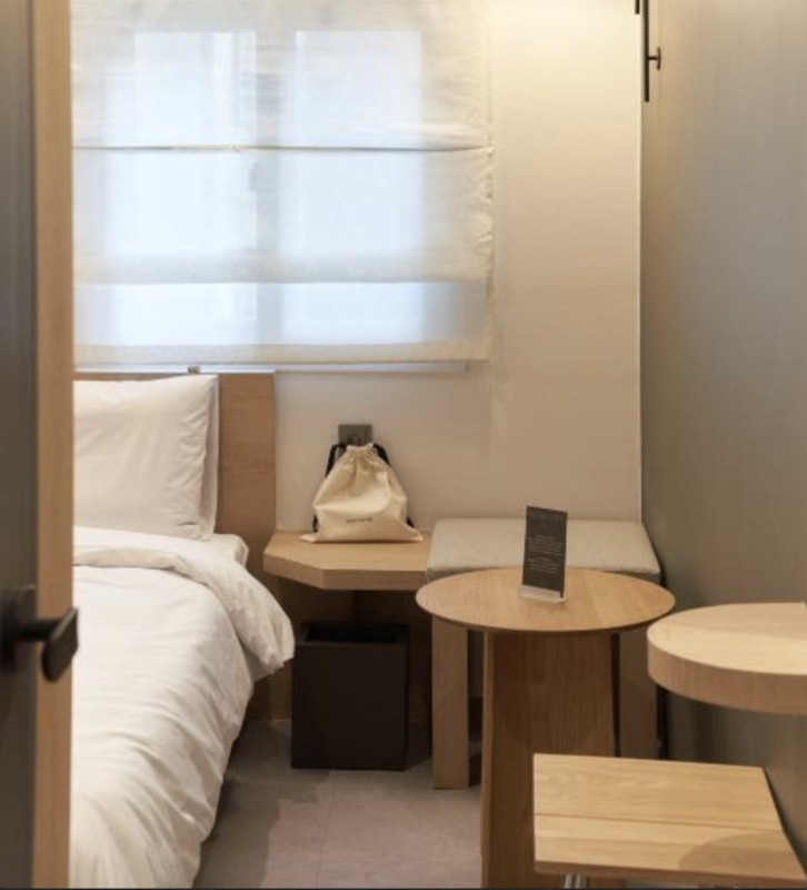 Compact hotel room corner with a single bed, white linens, a nightstand, and a round wooden table with a bag on it.