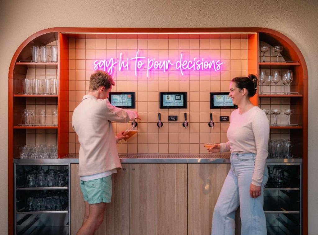 Two patrons at a neon-lit bar; a pink sign behind reads 'say hi to your decisions' as drinks are poured into glasses.