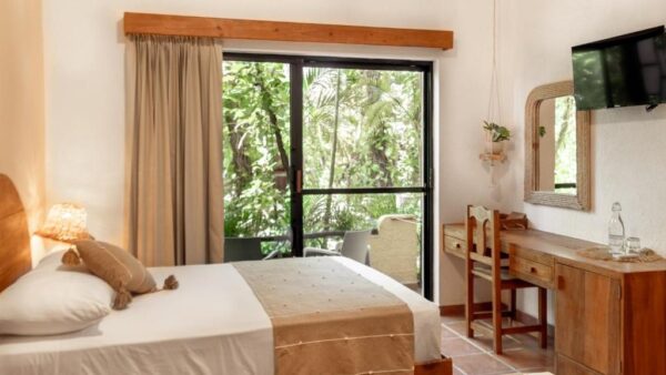 Hotel room with a neatly made bed, a sliding glass door to a lush balcony, and a wooden desk with a chair nearby.