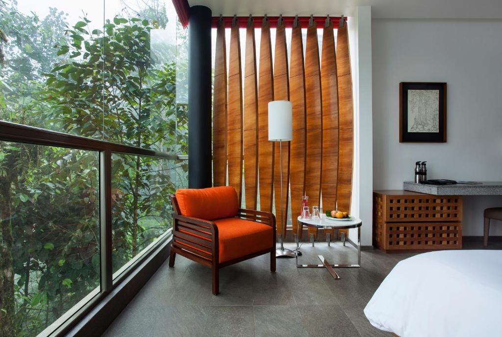Window-side hotel seating: orange armchair, round table with drinks, floor lamp, and a wooden slat divider.